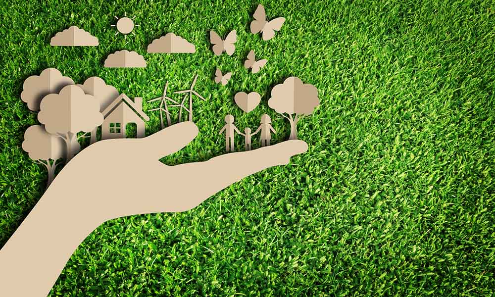 Α hand made of paper holding homes, people, trees and birds over a green grass background