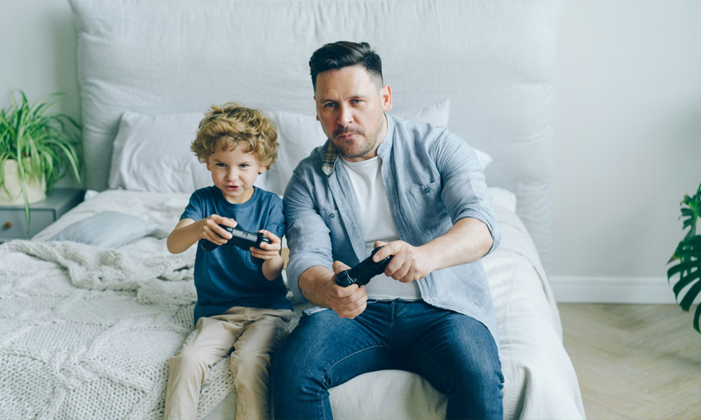 A father and his son playing videogames sitting on a white sheeted bed. 